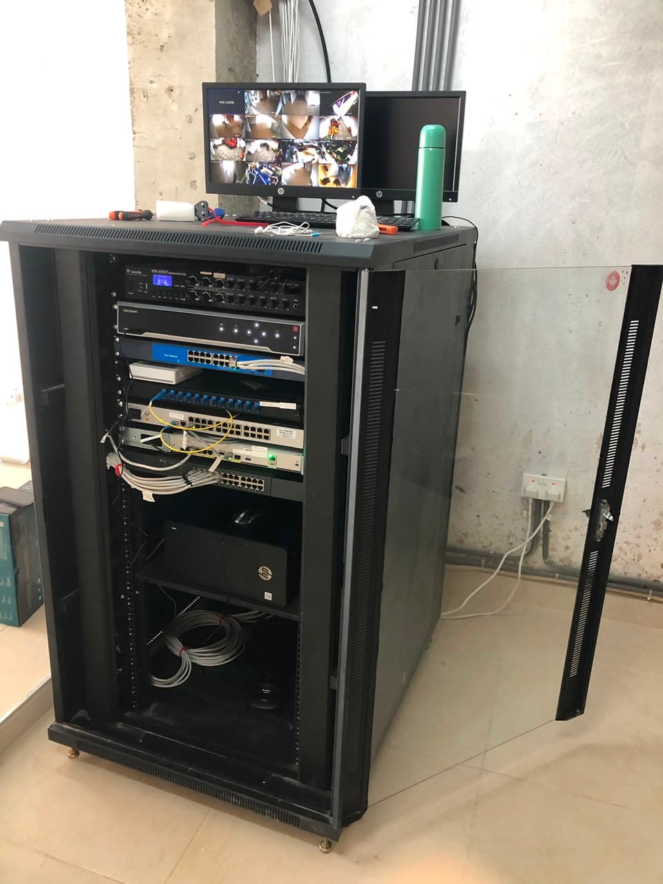 Floor-standing network cabinet containing a CCTV recorder, network switches and fibre patch panel, with a monitor showing a multi-camera live view